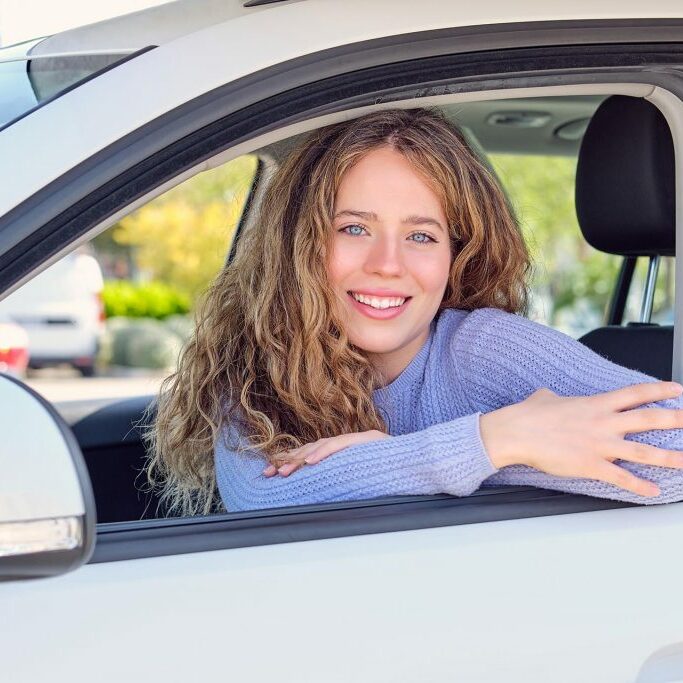 Cheerful young female driver in casual wear sitting in car parked in sunny city street and looking at camera with pretty smile